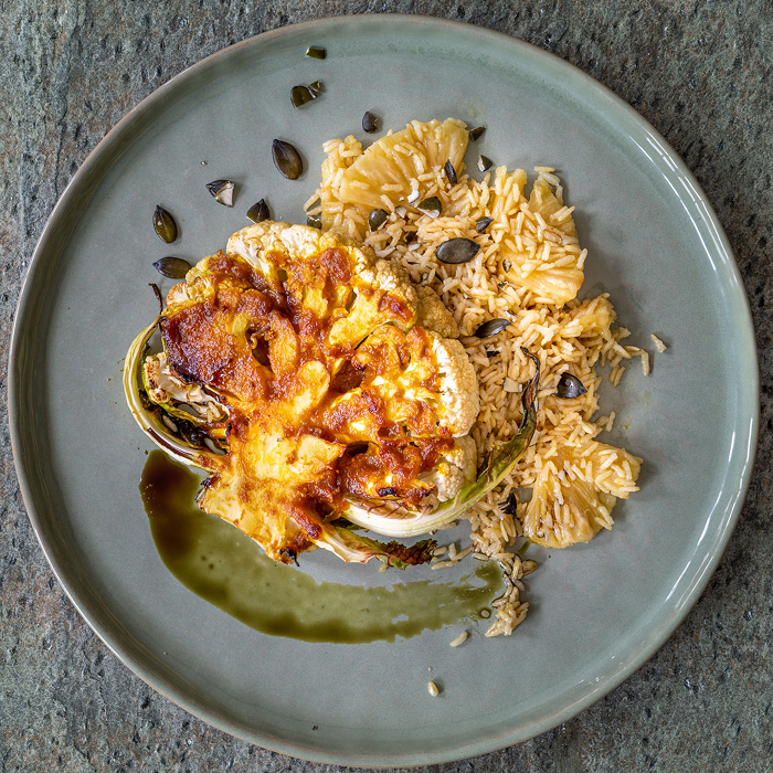 Ein geröstetes, goldbraun mariniertes Blumenkohl-Steak serviert mit lockerem Reis und Ananasstücken auf einem grauen Teller. Das Gericht ist mit dunklen Kernen bestreut und mit einem Streifen grüner Sauce garniert.